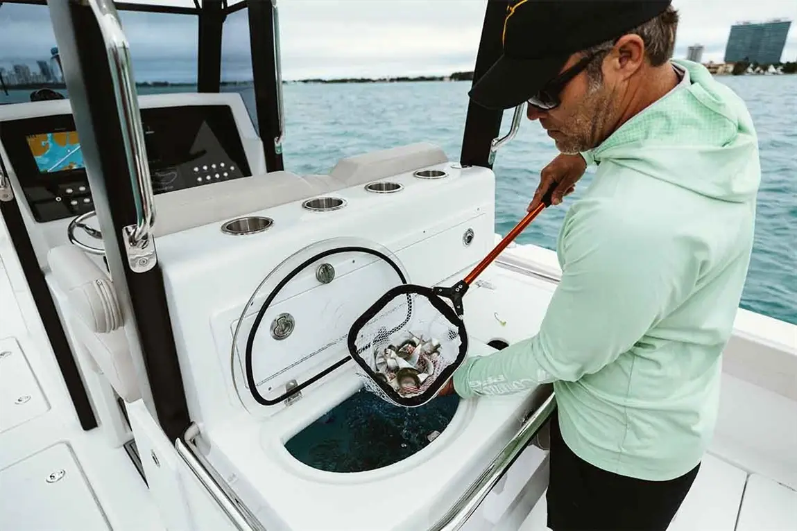 Man in a green shirt using a fishing net to catch fish from a blue water cooler on a boat.